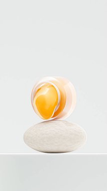 şeffaf krem kavanozu erimiş, yuvarlak bir taşın üzerine konmuş. Fotoğraf konseptinde beyaz zemin üzerinde kozmetik krem ürünü sergileniyor.