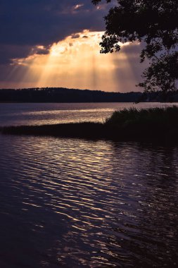 Batan güneş ışınları bulutların arasından görünüyor, gölün kıyısında ön planda.
