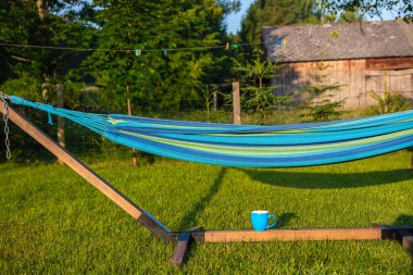 Mavi, ahşap hamak ve mavi kupa akşam güneşinin kırsal manzarasında yeşil çimlerin üzerinde duruyor.