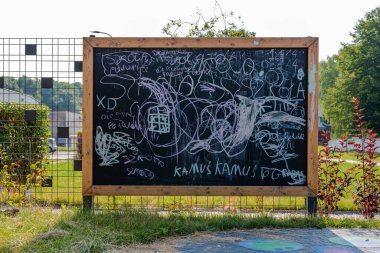 Oyun parkının dışına yerleştirilmiş büyük tahta tebeşir tahtası. Güneşli bir gün, arka planda ağaçlar.