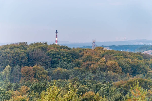 Mine shaft tower and factory chimney emerging form trees peaks, lightened by rising sun. Aerial view on autumn forest. Forest, factory and mining heap in the background. Katowice, Silesia, Poland.