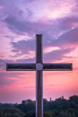 Tahta Hıristiyan haçı çelik bir çerçevede. Arka planda orman ve kilise var. Sabah ışığı, güzel, renkli, bulutlu gökyüzü. 