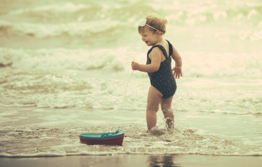 bir bebek kız sahilde arkasında bir tekne oyuncakla arıyorsunuz