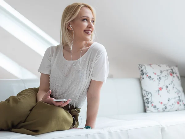 Mujer rubia sonriente en el sofá escuchando música en casa en el — Foto de Stock