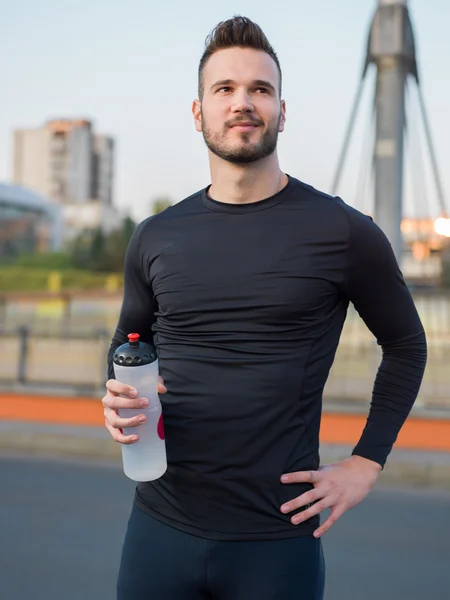 Joven corredor guapo con botella de agua sentado en un puente — Foto de Stock