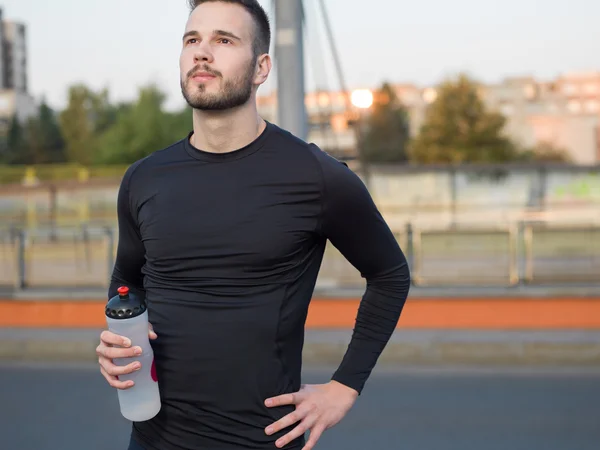 Joven corredor guapo con botella de agua sentado en un puente — Foto de Stock