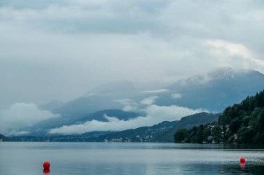 Millstaettersee Gölü kıyısından panoramik bir manzara. Göl yüksek dağlarla çevrilidir. Yüzeyde yüzen birkaç kırmızı şamandıra. Yukarıdaki yoğun ve karanlık bulutlar. Dinginlik ve sakinlik