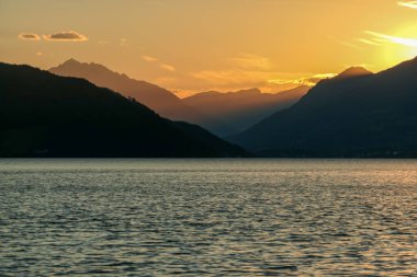 Avusturya 'da Millstaetter Gölü' nde gün batımı. Göl yüksek Alplerle çevrilidir. Gölün sakin yüzeyi güneş ışınlarını yansıtıyor. Güneş dağların arkasında batıyor. Biraz bulutlu. Doğal güzellik.