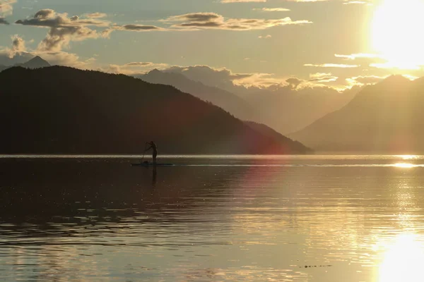 男はオーストリアのミルスターター湖の夕日の間にパドリングを立っています 湖は高いアルプスに囲まれています 陽射しを反射して湖の穏やかな表面 太陽は山の後ろに沈む — ストック写真