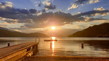 Millstaettersee Gölü 'ne giden ahşap bir iskele. Güneş yüksek Alpler 'in arkasında batıyor. Gölün sakin yüzeyi turuncu gökyüzünü ve dağları yansıtır. Gölde yüzen birkaç şamandıra var. Huzur