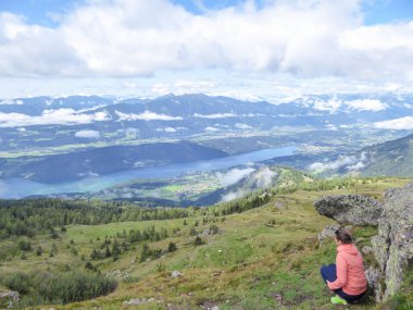Turuncu kapüşonlu bir kadın Avusturya Alplerinde Granattor 'dan Millstaetter Gölü manzarasının tadını çıkarıyor. Uzak göl yüksek dağlarla çevrilidir. Birkaç bulut yukarıda.