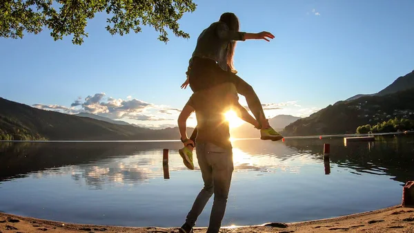 A man carrying a woman piggy back of Millstaetter lake during the sunset. The sun sets behind high Alps. Calm surface of the lake reflects the orange sky. They are having fun, playfulness. Day end