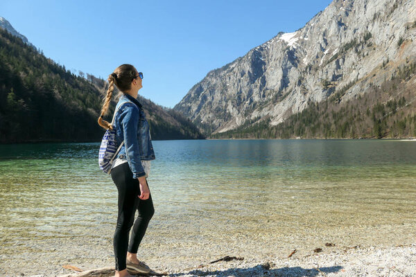 A woman walking along the shore of Leopoldsteiner lake in Austria. The lake is surrounded by high Alps. The shallow water is crystal clear, spring water has a calm surface. Early spring. Serenity