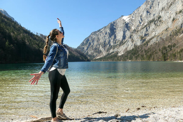 A woman spreading her arms wide open at the shore of Leopoldsteiner lake in Austria. The lake is surrounded by high Alps. The shallow water is crystal clear, spring water has a calm surface. Freedom