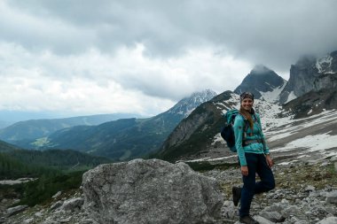 Avusturya 'nın Dachstein bölgesindeki yüksek Alplerde büyük bir kayanın üzerinde duran sırt çantalı bir kadın. Heyelan bölgesinden geçiyor. Uçsuz bucaksız vadideki manzaranın tadını çıkarıyor. Bulutlu. Özgürlük