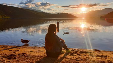 Millstaettersee sahilinde oturan kot ceketli bir kadın günbatımının tadını çıkarıyor. Etrafta yüzen birkaç ördek. Gölün sakin yüzeyi turuncu gökyüzünü ve dağları yansıtır. Yüksek Alpler.
