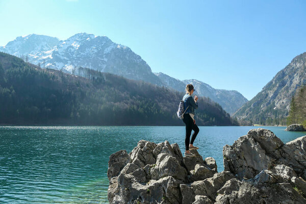 A woman with a backpack standing on a big stone at the shore of Leopoldsteinersee, an Alpine lake in Austria. There are high mountain chains around him. Crystal clear and calm surface. Discovering