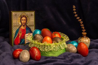 Renkli Paskalya yumurtaları, el yapımı güzel bir yuvada, arka planda bir Rus ikonu var. Küçük bir testide kurumuş fesleğen (cimum) filizi. 