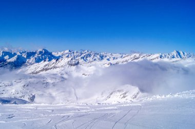 Dağlarda kar, uzakta sis, mavi gökyüzü, kayak pisti, karlı dağların manzarası.
