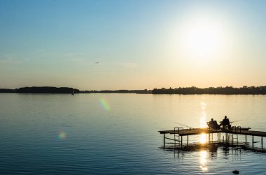 Rıhtımdaki balıkçılar balık yakalıyor. Masuria 'daki Swiecajty Gölü. Uzakta tekneler var. gün batımı.