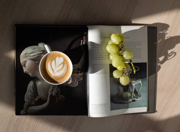 A cup of coffee with a leaf shaped design, on top of a magazine along with a group of grapes.