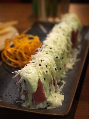 Yakın plan bir fotoğraf koyu dikdörtgen bir tabakta uzun bir suşi rulosu gösterir. Ekmek açık yeşil, kremalı sos, doğranmış beyaz garnitür (muhtemelen peynir veya daikon turpu) ve siyah susam çekirdekleri ile cömertçe kaplıdır. Rulonun içi...