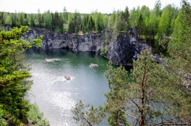 Mermer kayalarla çevrili yeşil göl suyu. Turistli tekneler göl boyunca yüzüyor. Uzun çam ağaçları kayalarda yetişir..