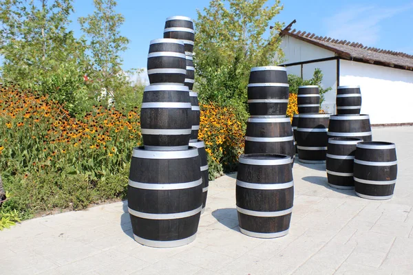There are dark oak barrels with metal hoops on the platform. Behind the barrels, a warehouse building, flowers are blooming. Bright blue sky.