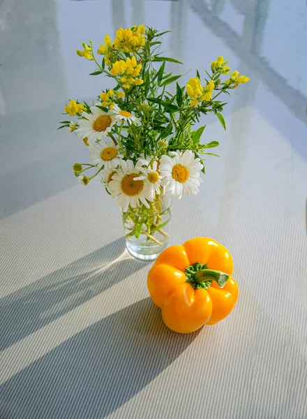Yellow peppers and a bouquet of chamomile and sweet pea flowers are illuminated by the sun and sit on a shiny gray tabletop. Reflection in the table top.