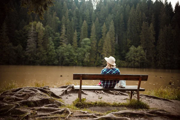 Rear view of a young woman in a denim shirt and hat, sitting on a bench next to a tree with many roots, admires the lake landscape, the pine forest and the relief of the mountains in the background, in Red Lake, Eastern Carpathians, Romania