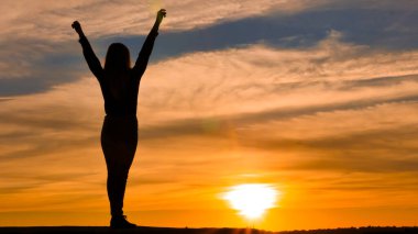 Kolları havada bir kadın silueti. Mutluluk ve neşe kavramı, turuncu bir günbatımının arka planında.