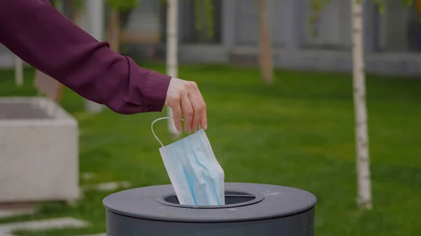 Woman hand throws a medical mask into the trash outdoors. The used face mask is dumped in a trash. Medical protective mask using to health protection from corona virus.