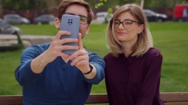 Gözlüklü iki arkadaş, güzel bir kadın ve yakışıklı bir adam dışarıda bankta oturup telefonla selfie çekiyorlar, mutlu bir şekilde gülümsüyorlar. Dışarıdaki sosyal ağlar. Portre. Kapatın. Yavaş çekim. 