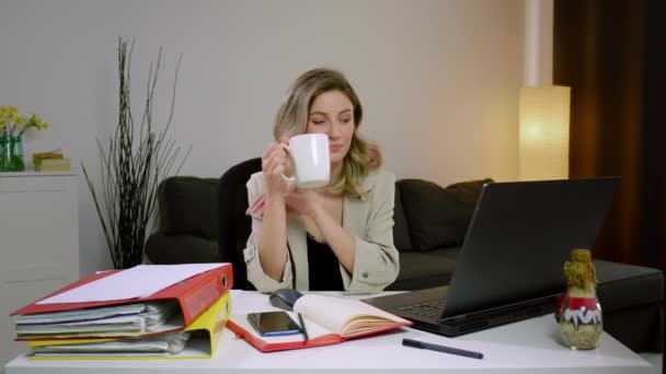 一位面带微笑的年轻女性在办公室的笔记本电脑上边喝咖啡边休息 并在工作暂停期间与一位朋友进行视频通话 休息一下上网工作 喝一杯咖啡 — 图库视频影像