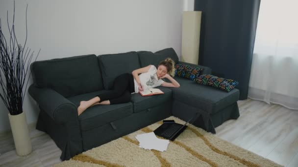 女学生躺在客厅的沙发上 在家学习 笔记本电脑在地板上 必要的文件夹 现代公寓视野开阔 大流行病期间在家进行的无聊研究 — 图库视频影像