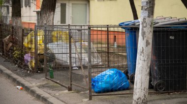 İki çöp konteyneri, ağacın yanında çöp poşeti ve plastik yığını, dışarıda güneş ışığı arka planında..