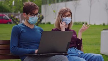 Koruyucu maskeli iki profesyonel iş arkadaşı arka planda bankta oturuyor. Dizüstü bilgisayarda çalışan gözlüklü bir adam. Cep telefonuyla konuşan bir kadın. Sağlık hizmetleri. Güvenlik önlemleri kavramı.