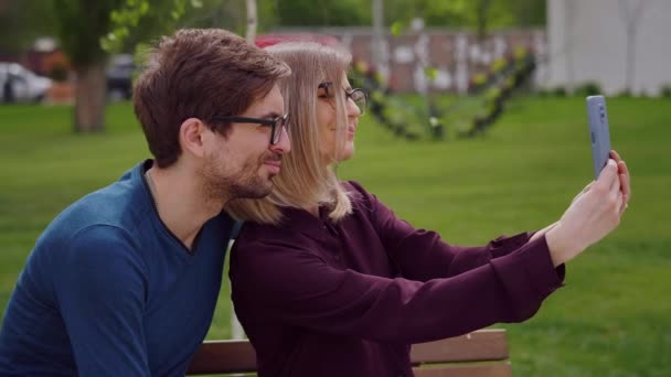 眼鏡をかけ きれいな女性とハンサムな男と2人の友人は 携帯電話で自撮りを取るの外のベンチに座って 幸せな笑顔を感じる スローモーション — ストック動画