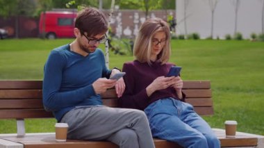 Açık hava parkında akıllı telefonları kullanan iki arkadaş, cep telefonu kullanan erkek ve kadın el ele mesajlaşarak alışverişe çıkıyor sosyal medyayı, mobil teknoloji yaşam tarzını ve bağımlılık kavramını kontrol ediyor. Kahve molası. Dışarıdaki sosyal ağlar.