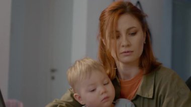 Genç bir anne, bebeğini kucağında tutarken dizüstü bilgisayarda birden fazla iş yapıyor. Yoğun bir çalışma alanının ortasındaki notlara ve görevlerine odaklanmış durumda..