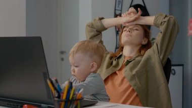 Anne evde çalışırken neşeli bebek, aile masasında rahat bir şekilde dizüstü bilgisayarını araştırıyor. Doğal ışık ve günlük iş hayatı dengesi ile.