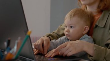 Anne dizüstü bilgisayarıyla bebeğini yazarken uzaktan iş ve çocuk bakımını dengeliyor. Ailesinin ve iş hayatının rahat bir ofis ortamında karışımını gösteriyor..