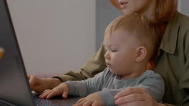 Meşgul bir anne, bebeği yanında oynarken dizüstü bilgisayarında çalışmasını dengeler ve ev ofisinin uzak çalışmalarının zorluklarını ve zevklerini gösterir..