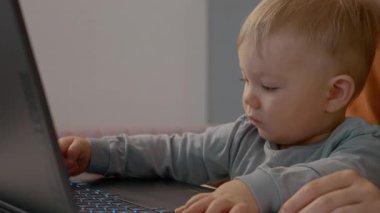 Bir bebek dizüstü bilgisayarın önünde oturur, klavyeye yazarken dikkatle odaklanır. Annesi yakınlarda, evlerindeki teknoloji keşiflerini destekliyor..