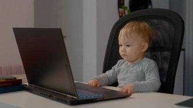 Sıcak bir ev ofisinde, bebek dizüstü bilgisayarın başına oturur, ekranı incelerken heyecan ve merak gösterir, teknolojiyle çocuk etkileşiminin hoş bir anını yaratır..