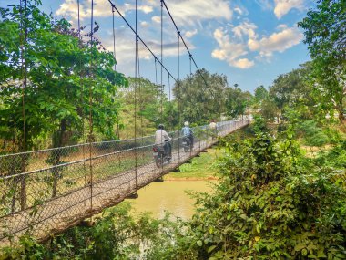 Üç Kamboçyalı motosikletçi dar, ahşap asma köprüsünün üzerinden geçiyor, miğfer takıyor, Kamboçya kırsalında bir nehri geçiyor..