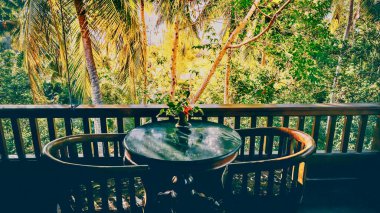 Yüksek kontrast sahnesi, iki kişilik bir masa ve gölgeli bir balkon, arka plandaki parlak tropikal ormana karşı siluet. Ubud, Bali.
