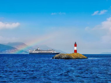 Arjantin 'in Ushuaia kenti yakınlarındaki Beagle Channel' da canlı kırmızı ve beyaz bantlarıyla Les Eclaireurs (yetenek avcıları) deniz fenerinin popüler turistik cazibesine sahip küçük bir ada görülüyor. Arka planda bir yolcu gemisi ve gökkuşağı var..