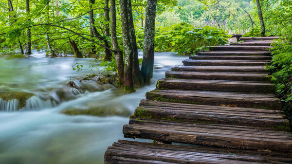 A long exposure image of water flowing beside a wooden boardwalk in the green forest of Plitvice Lakes National Park in Croatia.