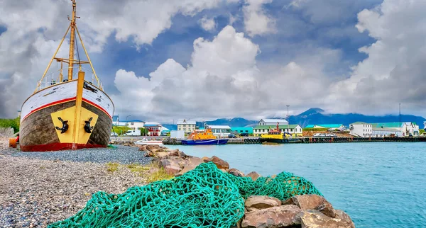 A commercial fishing harbor in a small coastal town in Iceland, with a green net in the foreground and an old wooden vessel out of the water and its keel buried in the ground up to its waterline to protect it from storms.